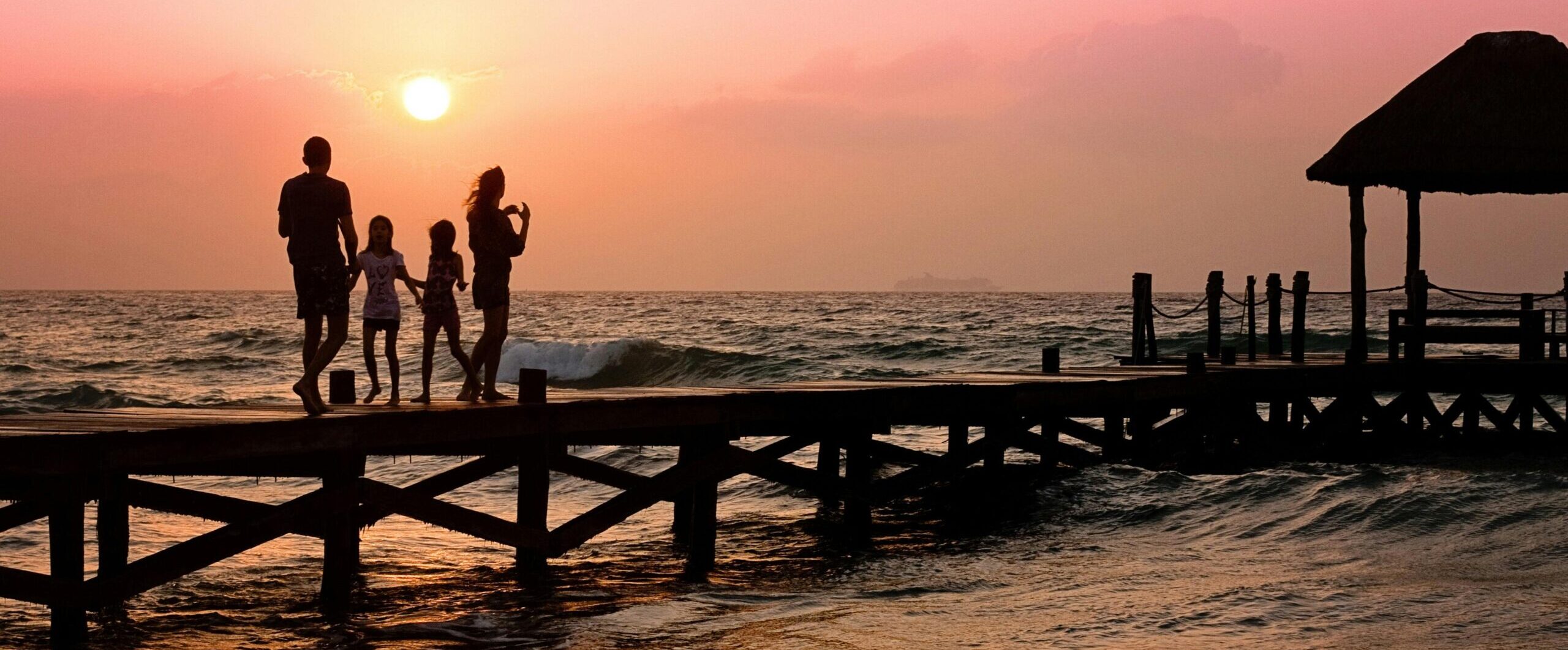 Silhouetted family enjoys a stroll on the beach pier at a vibrant sunset over the ocean waves.